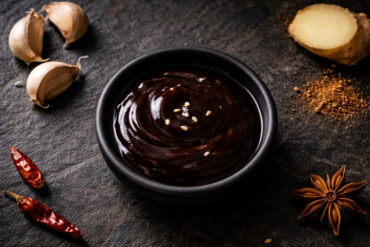 a small round matte black ceramic dish filled with thick, dark brown glossy hoisin sauce, sesame seeds scattered naturally on the surface of the sauce. Surrounding the dish whole garlic cloves with papery skin slightly peeling at the edges, one whole star anise, a thin diagonal slice of fresh ginger root showing pale yellow flesh at the cut edge, a small loose pinch of Chinese five-spice powder spilled directly onto the slate, and two small dried red chilies