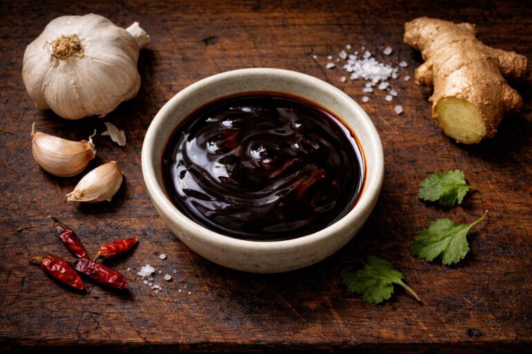 Top-down flat lay on a weathered dark teak cutting board with visible grain and knife marks. A small shallow ceramic bowl filled with dark, glossy oyster sauce. To the left: a whole garlic bulb with two cloves broken off and placed casually nearby, papery skin slightly peeling. To the right: a two-inch knob of fresh ginger root, unpeeled, with one rough cut end showing the pale yellow flesh. Scattered naturally around the bowl: four or five small dried red chilies, a pinch of coarse sea salt, and three or four cilantro leaves slightly wilted at the edges. natural window light from the upper left casting long soft shadows to the lower right.