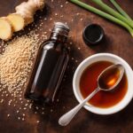 a small dark glass bottle of sesame oil, cap resting beside it, with a small white ceramic dish holding a spoonful of dark amber oil. Surrounding props: a loose scatter of raw sesame seeds directly on the surface, a thin diagonal slice of fresh ginger root, two trimmed green onion stalks