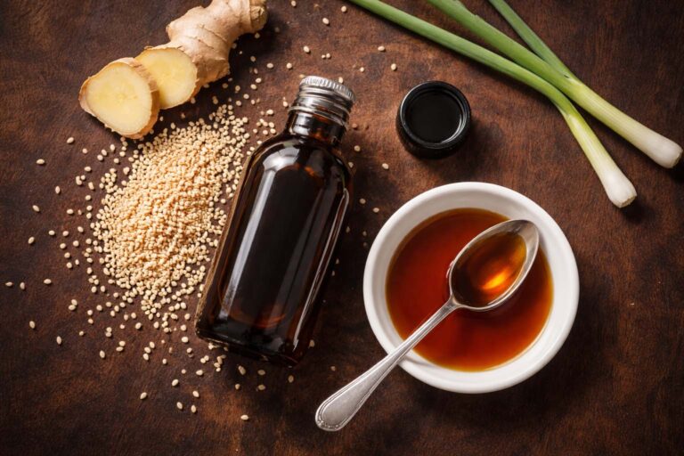 a small dark glass bottle of sesame oil, cap resting beside it, with a small white ceramic dish holding a spoonful of dark amber oil. Surrounding props: a loose scatter of raw sesame seeds directly on the surface, a thin diagonal slice of fresh ginger root, two trimmed green onion stalks