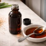 a small dark glass bottle of sesame oil, cap beside it, and a white ceramic dish with a spoonful of dark amber oil. Right side, slightly out of focus: the middle half of a white refrigerator door slightly ajar