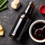 an open bottle of teriyaki sauce with the cap resting beside it, a small white ceramic dish with a spoonful of dark glossy sauce poured into it.
