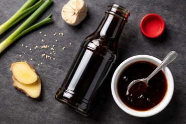 an open bottle of teriyaki sauce with the cap resting beside it, a small white ceramic dish with a spoonful of dark glossy sauce poured into it.