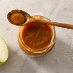 An open glass jar of caramel sauce with a wooden spoon placed on top. Left: Half a green apple. Right: A small pinch of flaky sea salt directly onto the marble.