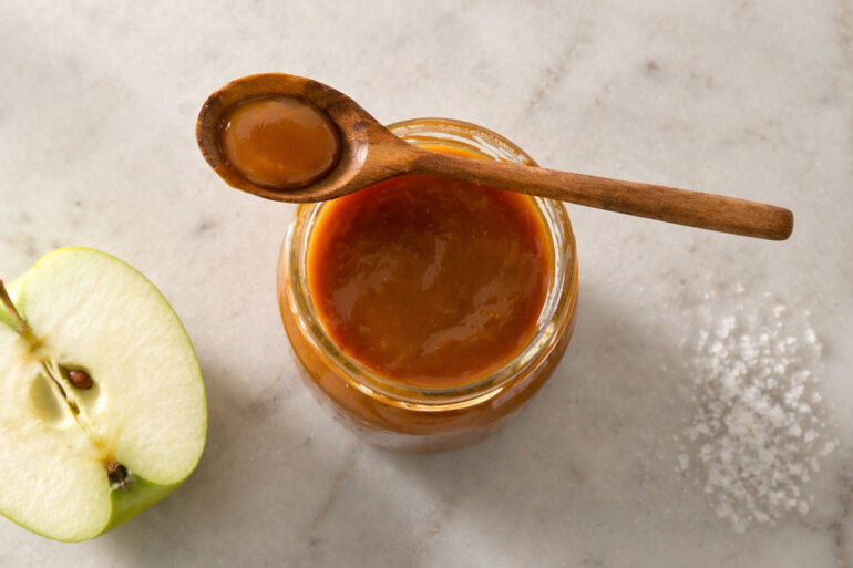 an open glass jar of caramel sauce with a wooden spoon resting across the top. Left: a halved green apple. Right: a small pinch of flaky sea salt directly on the marble.