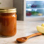Kitchen counter, warm natural light. Left foreground: a sealed jar of caramel sauce, no readable label, amber sauce visible through glass. background slightly out of focus: open refrigerator door with cool interior light. On the counter: a wooden spoon and two apple slices