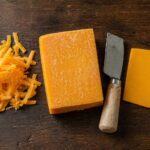 Flat lay on a dark wood surface. Center: a block of aged sharp cheddar with visible white spots on the surface and a cheese knife resting alongside. Left: a small pile of freshly shredded cheddar.