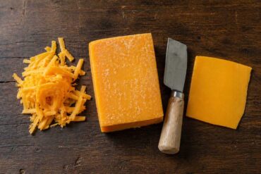 Flat lay on a dark wood surface. Center: a block of aged sharp cheddar with visible white spots on the surface and a cheese knife resting alongside. Left: a small pile of freshly shredded cheddar.
