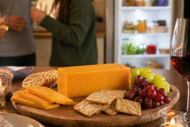 A cheese board scene at a dinner party. A block of cheddar sits on a wooden board with crackers and grapes. A soft-focus refrigerator is visible in the background.