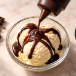 a squeeze bottle of chocolate syrup mid-drizzle over a glass bowl of vanilla ice cream. Left: a small square of dark chocolate broken in two. Right: a clean tablespoon.