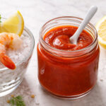 An open glass jar of cocktail sauce, lid closed, small ceramic spoon placed inside. Left: Three or four large cooked prawns arranged naturally on a small white plate. Right: Showing the pulp of half a lemon. Scatter props directly on the surface: a few small capers, a pinch of coarse salt, a small sprig of fresh dill or parsley. The sauce is bright red and shiny