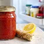 one sealed jar of cocktail sauce, no readable label, bright red sauce visible through glass. Right side slightly out of focus: a refrigerator door open, cool interior light visible, condiment shelf with a few other jars