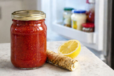 one sealed jar of cocktail sauce, no readable label, bright red sauce visible through glass. Right side slightly out of focus: a refrigerator door open, cool interior light visible, condiment shelf with a few other jars