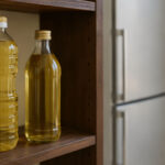 Kitchen pantry shelf in soft natural light. Two bottles of cooking oil on a dark wooden shelf, a closed refrigerator door visible in the background slightly out of focus.