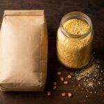 Flat lay on a dark wood surface. Two containers side by side: left, a sealed paper bag of standard degerminated cornmeal; right, a glass jar of coarser stone-ground cornmeal with some spilled grains. A few dried yellow corn kernels scattered nearby