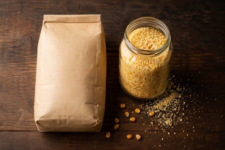 Flat lay on a dark wood surface. Two containers side by side: left, a sealed paper bag of standard degerminated cornmeal; right, a glass jar of coarser stone-ground cornmeal with some spilled grains. A few dried yellow corn kernels scattered nearby