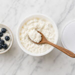 an open container of cottage cheese with a wooden spoon resting across it, curds visible. Left: a small bowl of the same with fresh blueberries. Right: the container lid with a visible date stamp