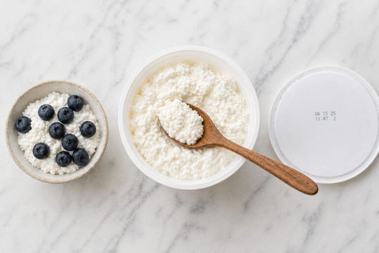 an open container of cottage cheese with a wooden spoon resting across it, curds visible. Left: a small bowl of the same with fresh blueberries. Right: the container lid with a visible date stamp