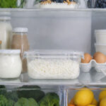Interior refrigerator shot. A clear sealed container of cottage cheese sits prominently on a back shelf alongside other fresh dairy items. The fridge is clean and well-organized