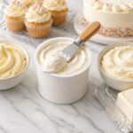 Clean flat lay on a white marble surface. Center: an open can of store-bought frosting with a spatula resting on it. Left: a small bowl of homemade buttercream with a swirl on top. Right: a bowl of cream cheese frosting next to a block of cream cheese