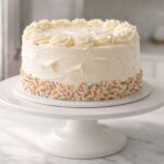 A frosted two-layer cake on a white cake stand sits on a kitchen counter. Behind it, slightly out of focus, a refrigerator door is partially open. The cake has visible swirls of buttercream