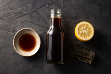 Flat lay on a dark slate surface. Center: a bottle of Kikkoman ponzu with the cap off. Left: a small ceramic dipping bowl with ponzu. Right: a halved yuzu and a small piece of kombu.