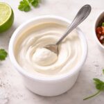 an open white plastic container of sour cream with a clean silver spoon resting inside. Left: a lime cut in half. Right: a small bowl of fresh salsa. Scattered props: a few cilantro leaves and a pinch of coarse salt directly on the surface