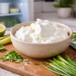 Looking a bit into an open refrigerator. Foreground: sour cream and chives with lemon on a cutting board. The other dairy items behind it are slightly out of focus.