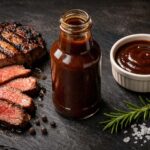 one open bottle of steak sauce, no readable label, deep brown sauce visible. Left: a thick-cut grilled steak, seared and resting, sliced to show the interior. Right: a small ramekin with a pool of steak sauce. Scattered props directly on the surface: a few whole black peppercorns, a sprig of fresh rosemary, coarse sea salt.