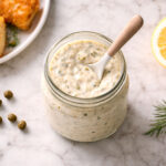 one open glass jar of tartar sauce with a small ceramic spoon resting in it. Left: two or three pieces of golden fried fish on a small white plate. Right: a halved lemon showing the flesh. Scattered props: a few capers and a small sprig of fresh dill directly on the surface