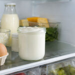 Interior refrigerator shot. A sealed tub of Greek yogurt sits prominently on a back main shelf, not the door. Clean, organized fridge with other dairy items visible.