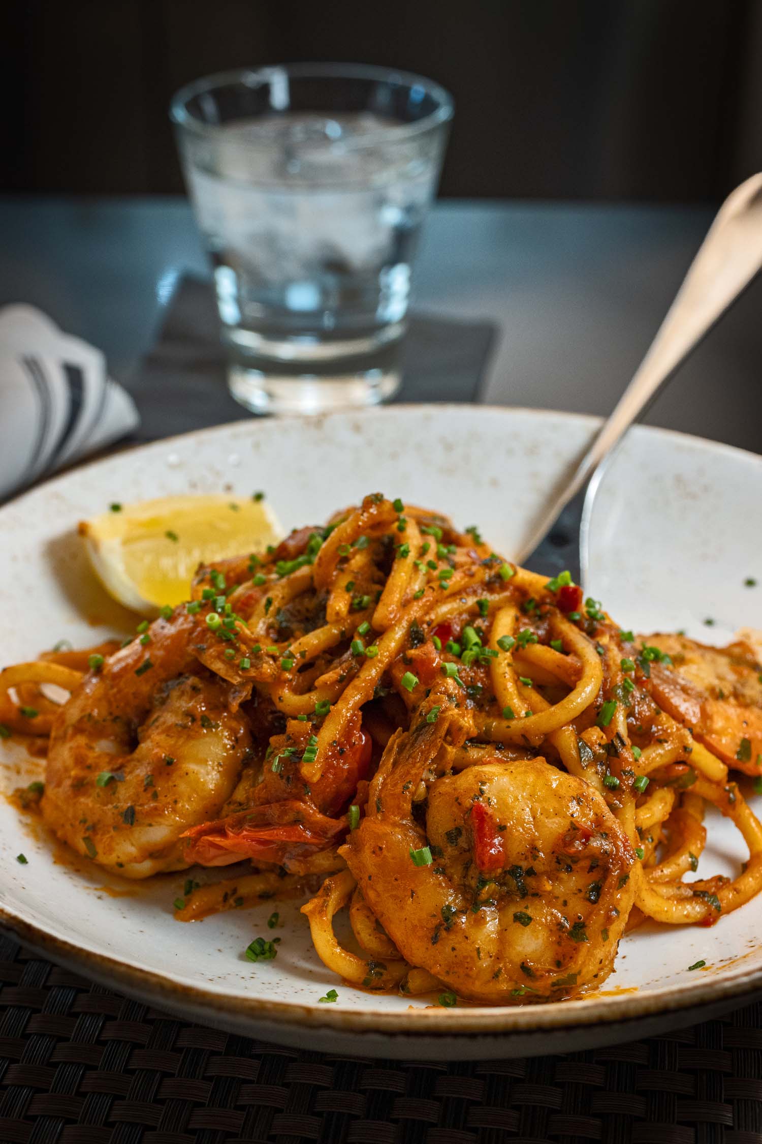 A generous serving of Spicy Shrimp Bucatini at Fireclay Restaurant at Hotel Washington DC, presented on a speckled white ceramic plate, featuring thick bucatini pasta coated in a deep orange-red spicy tomato broth, topped with several large whole shrimp, finely chopped green chives and fresh herbs, a wedge of lemon on the side, and a silver fork resting in the dish, with a glass of ice water and folded striped napkin visible in the soft-focus background