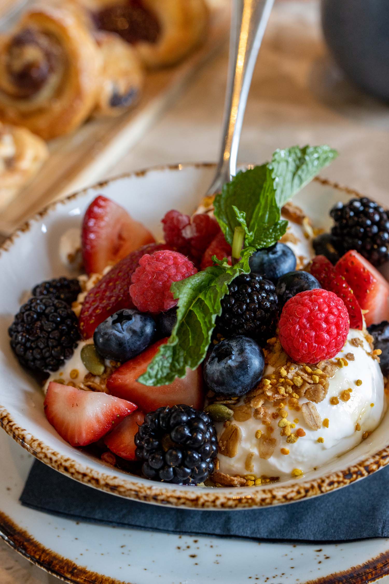 A close-up of the Mixed Berry Yogurt Bowl served at breakfast at Fireclay Restaurant at Hotel Washington DC, showing a rustic brown-rimmed ceramic bowl filled with creamy white Greek yogurt topped with a generous arrangement of fresh blackberries, blueberries, sliced strawberries, and raspberries, sprinkled with golden bee pollen, house-made granola, and pumpkin seeds, garnished with a fresh mint sprig, with a silver spoon inserted in the bowl and a wooden board of warm pastries including a chocolate croissant visible in the soft-focus background