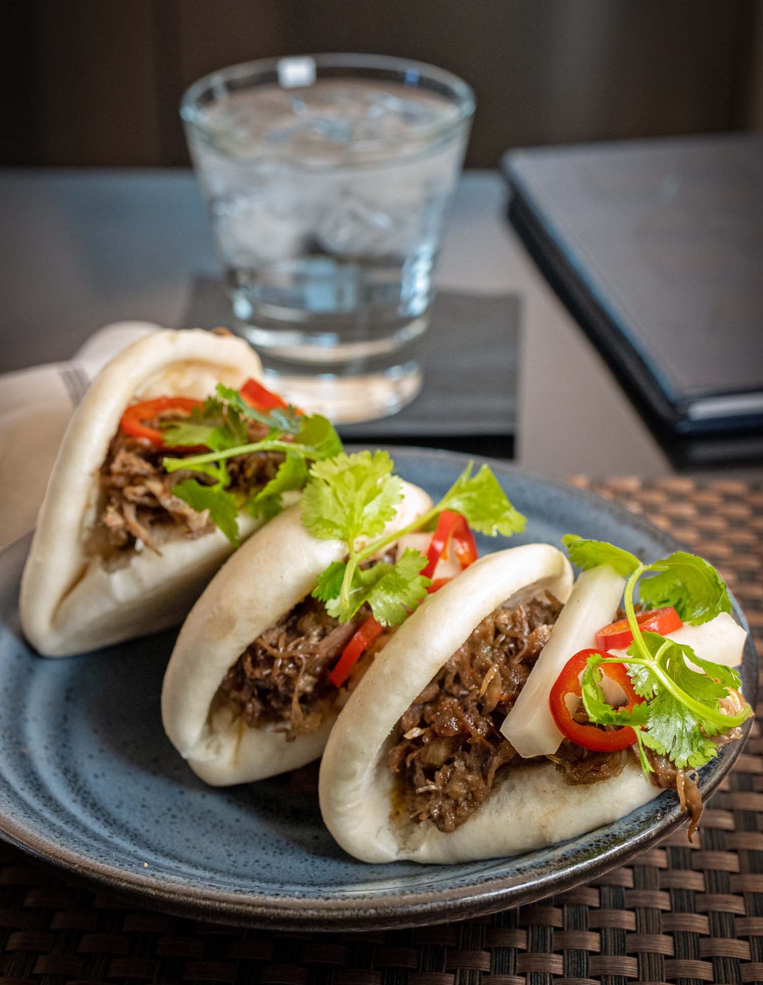 Three steamed Duck Confit Bao Buns served on a slate blue ceramic plate at Fireclay Lobby Bar at Hotel Washington DC, each soft white bao bun generously filled with tender shredded duck confit, topped with sliced red chili peppers, fresh cilantro leaves, and pickled accompaniments, presented on a woven dark rattan surface with a rocks glass of ice water and a small black check presenter visible in the soft-focus background