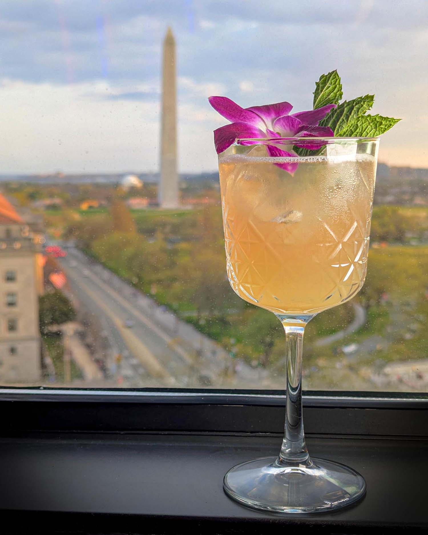The Power to the Peaches signature cocktail at VUE Rooftop at Hotel Washington DC, served in a crystal cut wine glass garnished with a purple orchid and fresh mint sprig, photographed on a black window ledge with the Washington Monument and National Mall visible in soft focus in the background at dusk