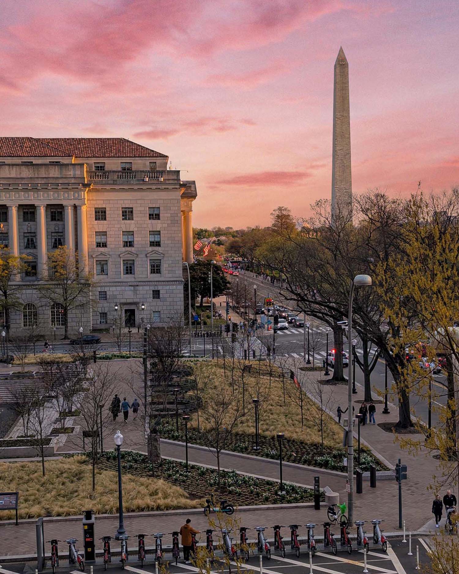 The view from VUE- A wide aerial view of Washington DC at sunset showing the Washington Monument silhouetted against a vivid pink and coral sky, with a neoclassical federal building lit in warm golden light to the left, Pennsylvania Avenue stretching into the distance lined with flags and cherry blossom trees, Capital Bikeshare docking stations in the foreground, and pedestrians and cyclists visible at street level