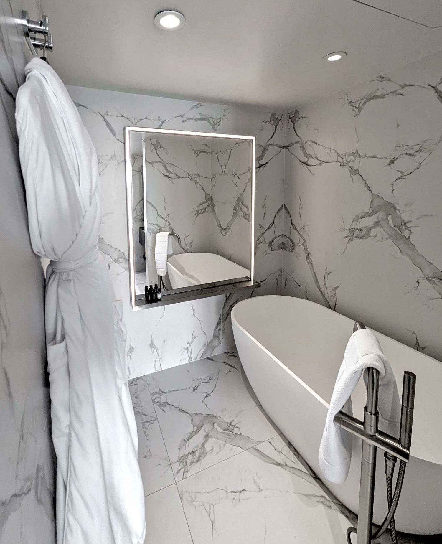 The full marble bathroom of the Monument View King Spa Suite at Hotel Washington DC, featuring floor-to-ceiling white Carrara-style marble with dramatic grey veining on all walls and floors, a sculptural freestanding white soaking tub positioned in the corner with a chrome freestanding tub filler and handheld shower attachment, a white towel draped over the chrome fixture, a square backlit LED mirror mounted on the marble wall with a small shelf below holding dark glass toiletry bottles, and a plush white hotel bathrobe hanging on a chrome hook on the left wall