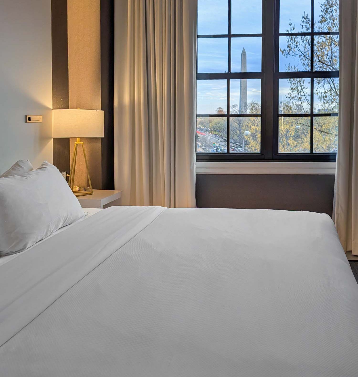 The king bed of the Monument View King Spa Suite at Hotel Washington DC photographed from the foot of the bed, showing crisp white Egyptian cotton linens, a warm bedside lamp with a geometric gold tripod base, floor-length cream curtains partially open, and a large multi-pane black-framed window with the Washington Monument visible in the distance against a soft blue sky with bare spring trees in the foreground