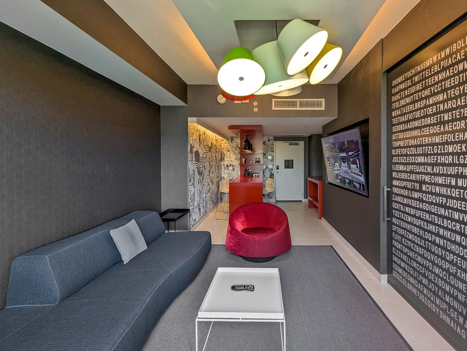 The living room of the Monument View King Spa Suite at Hotel Washington DC, featuring a gray sectional sofa, bold red lounge chair, wall-sized flat screen television, colorful pendant light fixture in green and white, and a black wall covered in white text lettering, with a red lacquered bar and Amit Greenberg mural visible in the background