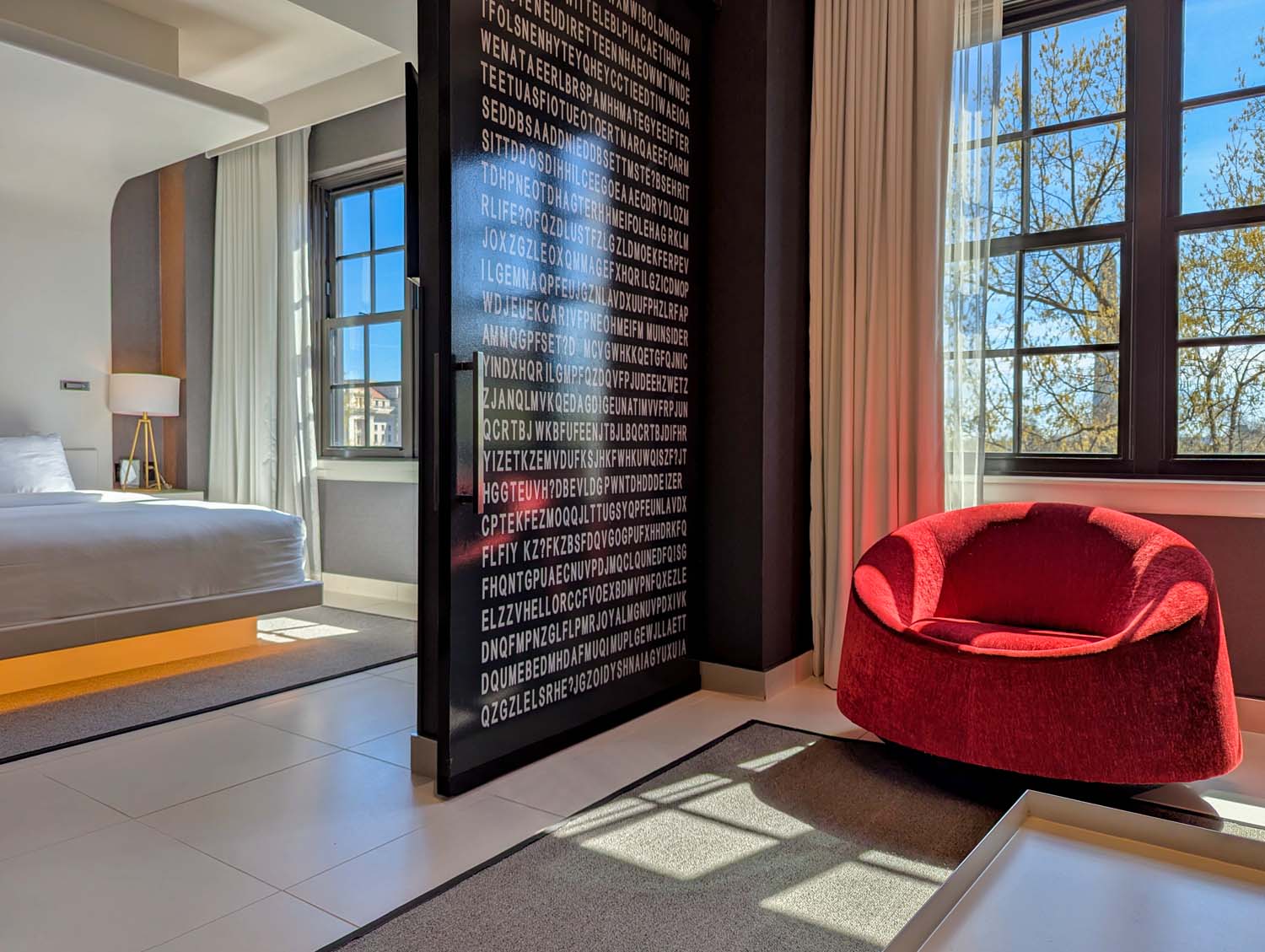 The bedroom area of the Monument View King Spa Suite at Hotel Washington DC, showing a king bed with white linens and warm amber accent lighting, a large black door panel covered floor to ceiling in white lettering, a bold red velvet lounge chair, and floor-to-ceiling windows letting in bright natural light