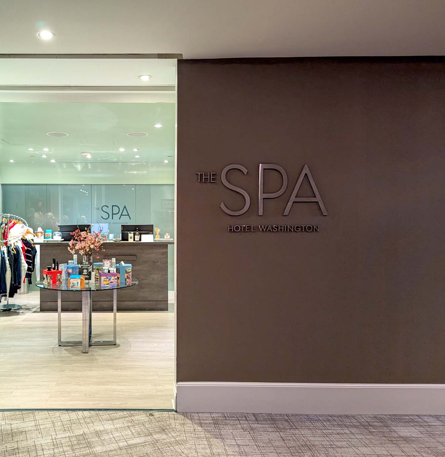 The entrance to The Spa at Hotel Washington DC, showing the spa retail area through glass doors on the left with cherry blossom décor and skincare products on display, and a large dimensional sign reading THE SPA HOTEL WASHINGTON mounted on a warm brown wall to the right