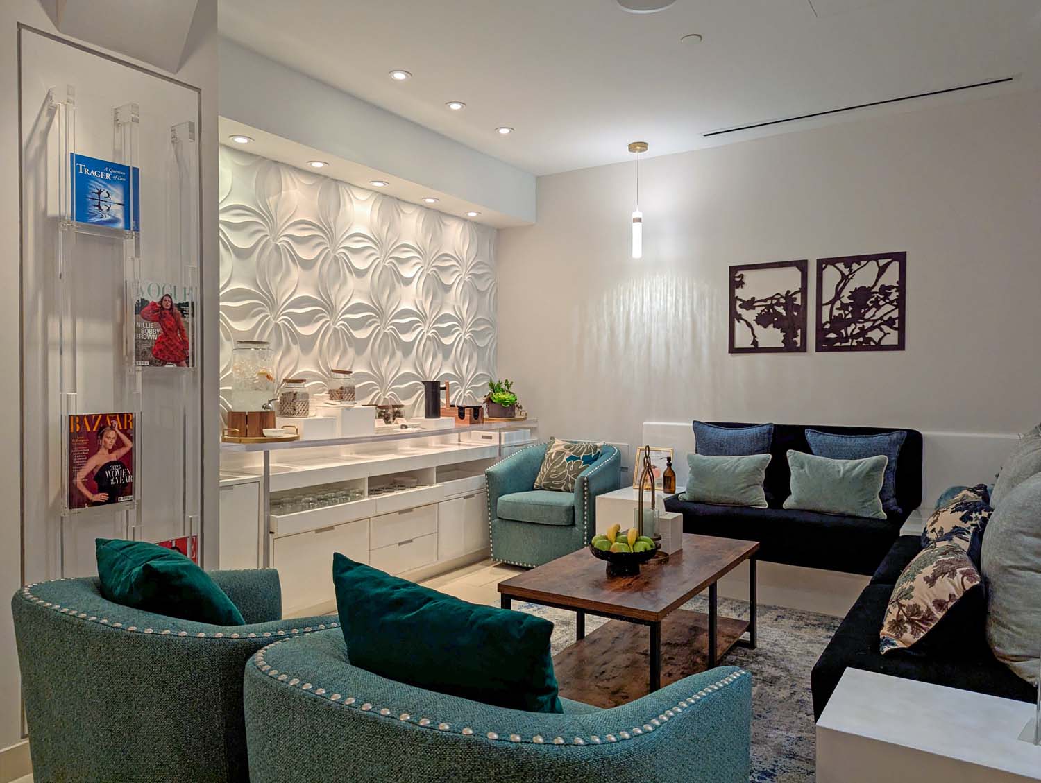 The women's relaxation lounge at The Spa at Hotel Washington DC, featuring teal velvet curved armchairs with nail-head trim, a dark navy sofa with teal and floral pillows, a rustic wood and metal coffee table with a bowl of lemons, a white sculptural 3D floral accent wall with backlit shelving, an acrylic magazine display rack, and two dark metal cherry blossom silhouette artworks on the wall