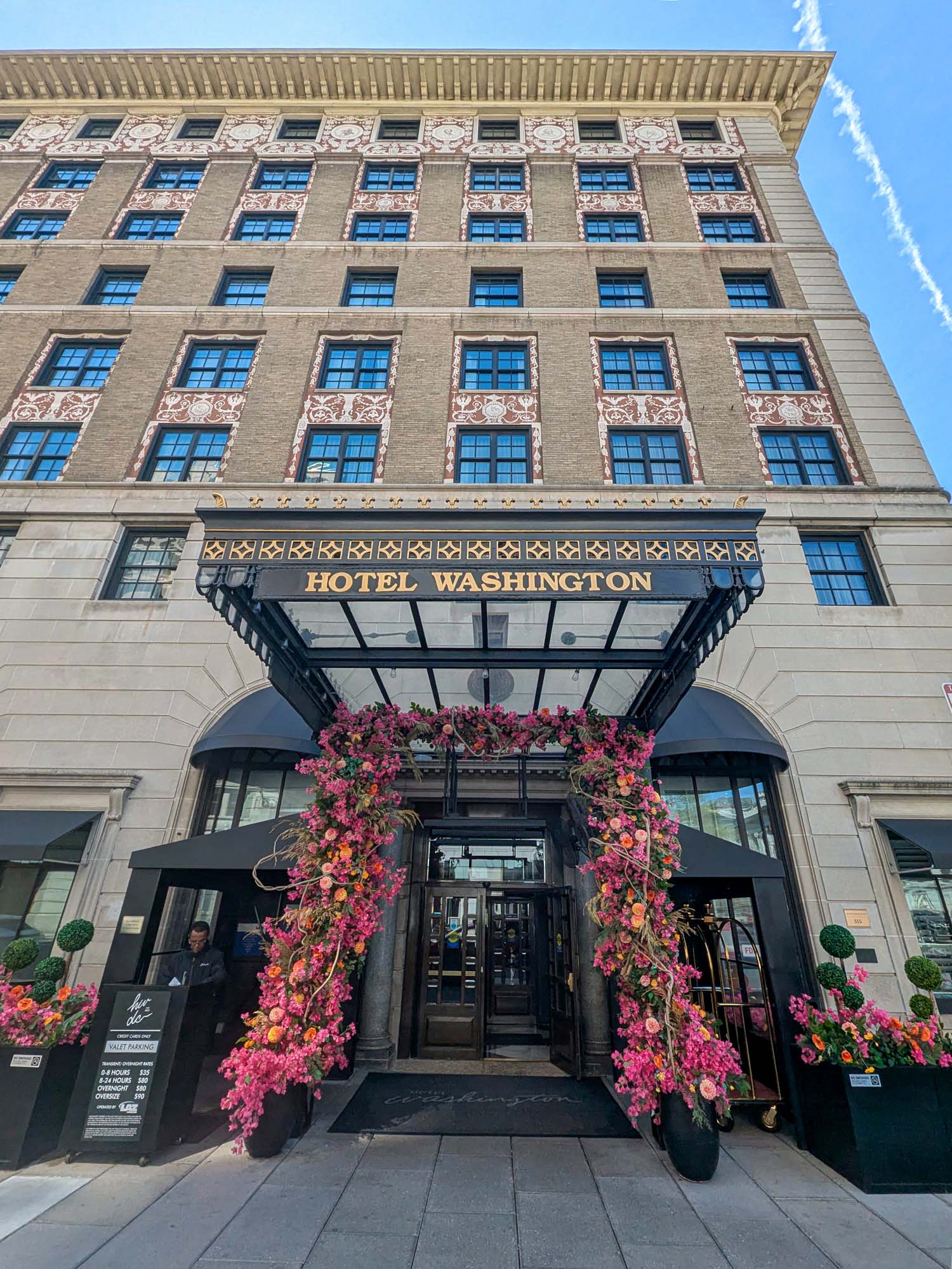 The Hotel Washington DC facade photographed from street level looking up during cherry blossom season, showing the full height of the historic brick building with ornate terracotta decorative panels, the black entrance canopy with Hotel Washington in gold letters, and an elaborate floral arch of pink and orange blooms flanked by topiary balls and potted flowers