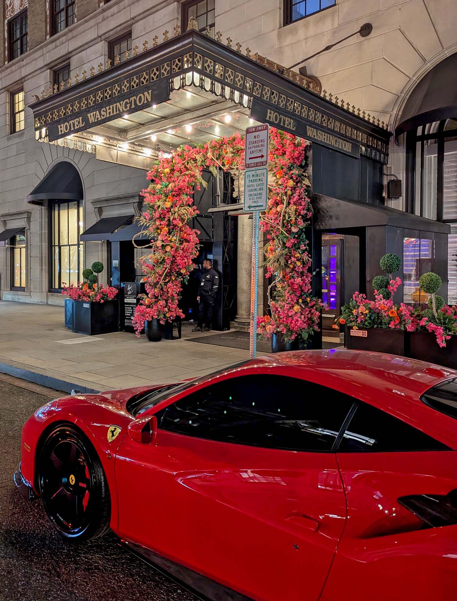 The Hotel Washington DC entrance lit up at night during cherry blossom season, with dramatic pink and orange floral arch installations framing the black entrance canopy displaying the Hotel Washington name in gold lettering, topiary plants flanking the doorway, a doorman standing at the entrance, and the front hood of a red Ferrari visible in the foreground