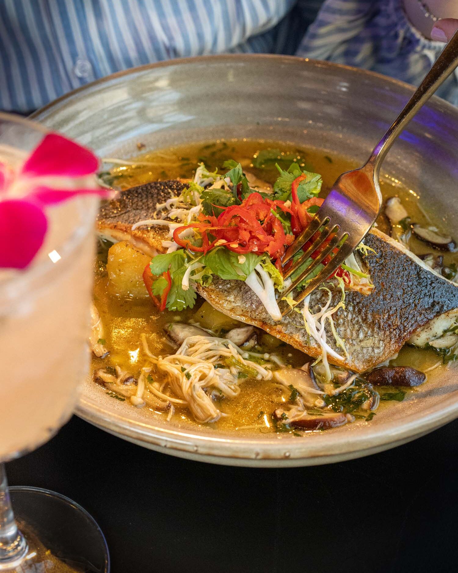 Crispy Branzino served at VUE Rooftop at Hotel Washington DC, featuring two seared fish fillets with crackling skin presented in a shallow golden broth with enoki mushrooms, wilted greens, and potato, topped with bright red pickled pepper strips and fresh cilantro, with a pink cocktail flower visible at the left edge of the frame