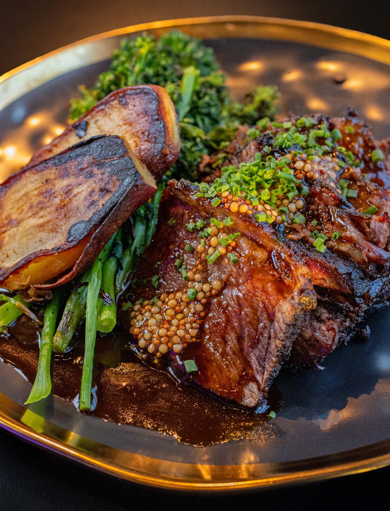 The 48-Hour Smoked Short Rib served at VUE Rooftop at Hotel Washington DC, presented on a dark plate with a gold rim, showing sliced deeply glazed and smoked beef short rib topped with pickled mustard seeds and chopped chives, alongside charred white sweet potato and broccolini in a rich dark pan sauce