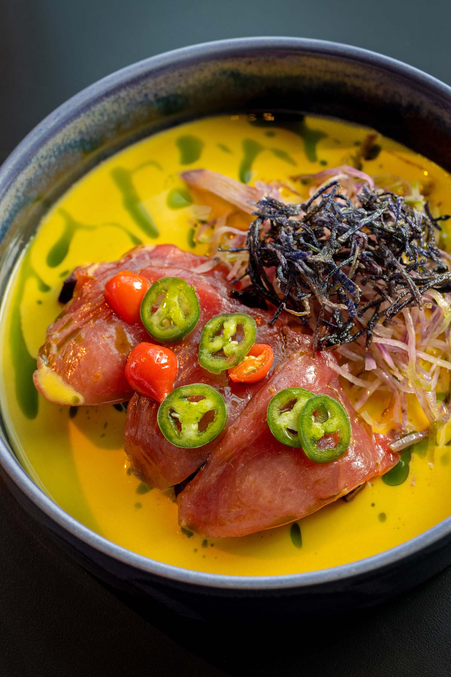 Yellowfin Tuna Crudo served at VUE Rooftop at Hotel Washington DC, presented in a deep blue ceramic bowl filled with vivid golden yellow sauce, topped with thick slices of raw tuna, sliced green jalapeños, small red cherry peppers, pickled shallots, and a nest of crispy fried dark seaweed, finished with green herb oil drizzled over the sauce