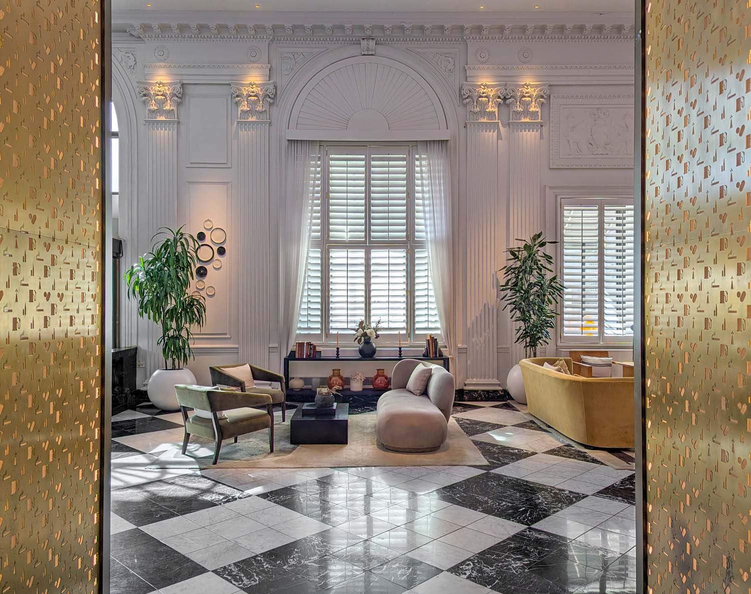 The Hotel Washington DC lobby lounge viewed through ornate gold perforated metal panels, revealing an elegant cream and white room with soaring plaster coffered ceilings featuring Corinthian columns and an arched fanlight window, black and white diamond marble floors, a curved dove gray sofa on a cream rug, olive green and mustard curved accent chairs, tall tropical plants, and decorative circular wall sculptures