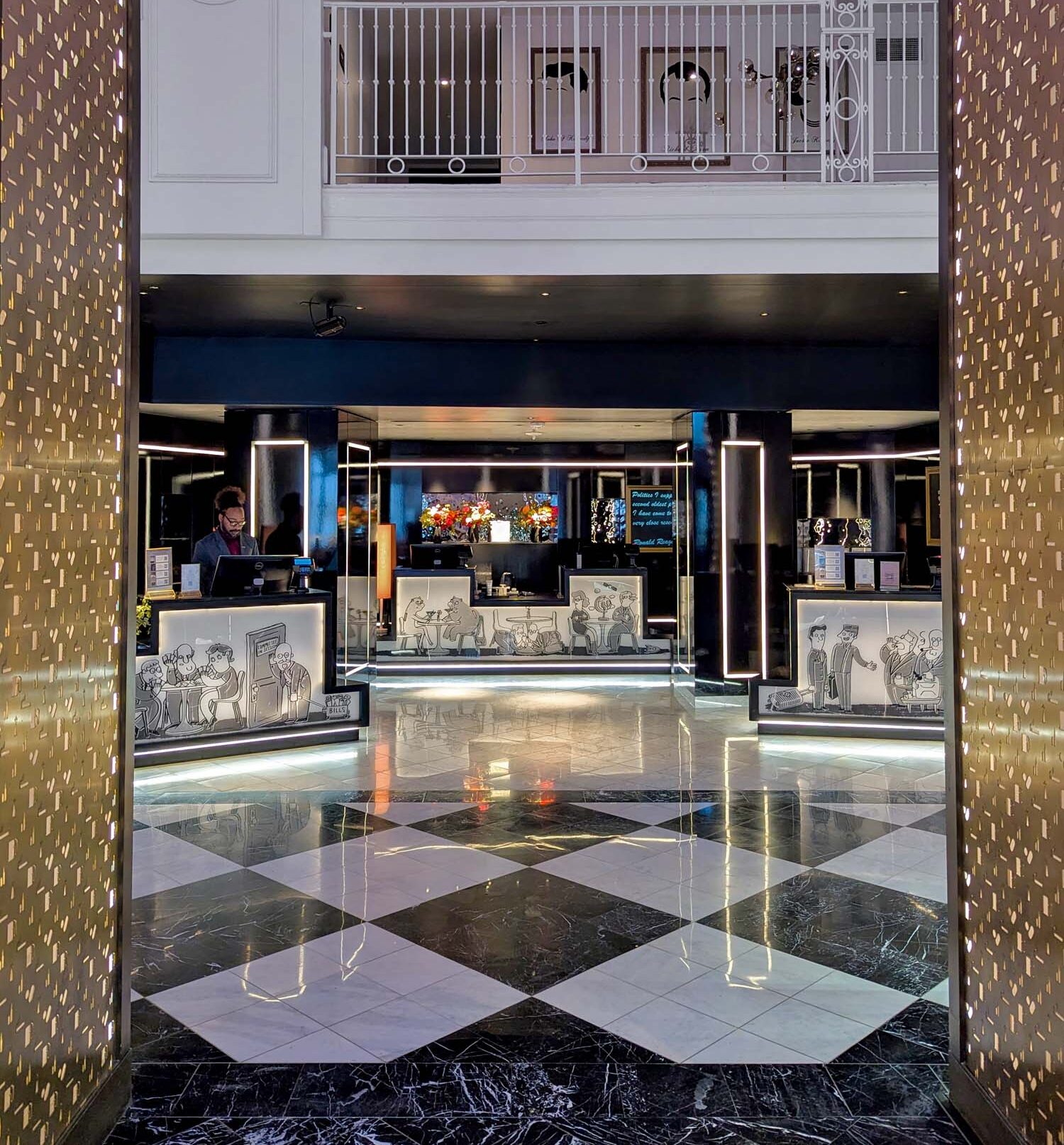 The Hotel Washington DC check-in area viewed through the gold brass perforated entrance panels, showing the dramatic black and white diamond marble floors, illuminated front desk stations with custom black and white cartoon illustration panels depicting Washington DC political scenes, black neon light column accents, a staff member at the desk, and a second floor mezzanine with white iron railings visible above