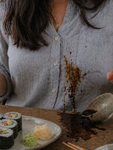 A woman eating sushi and splashing soy sauce on her shirt as the bowl is tipped